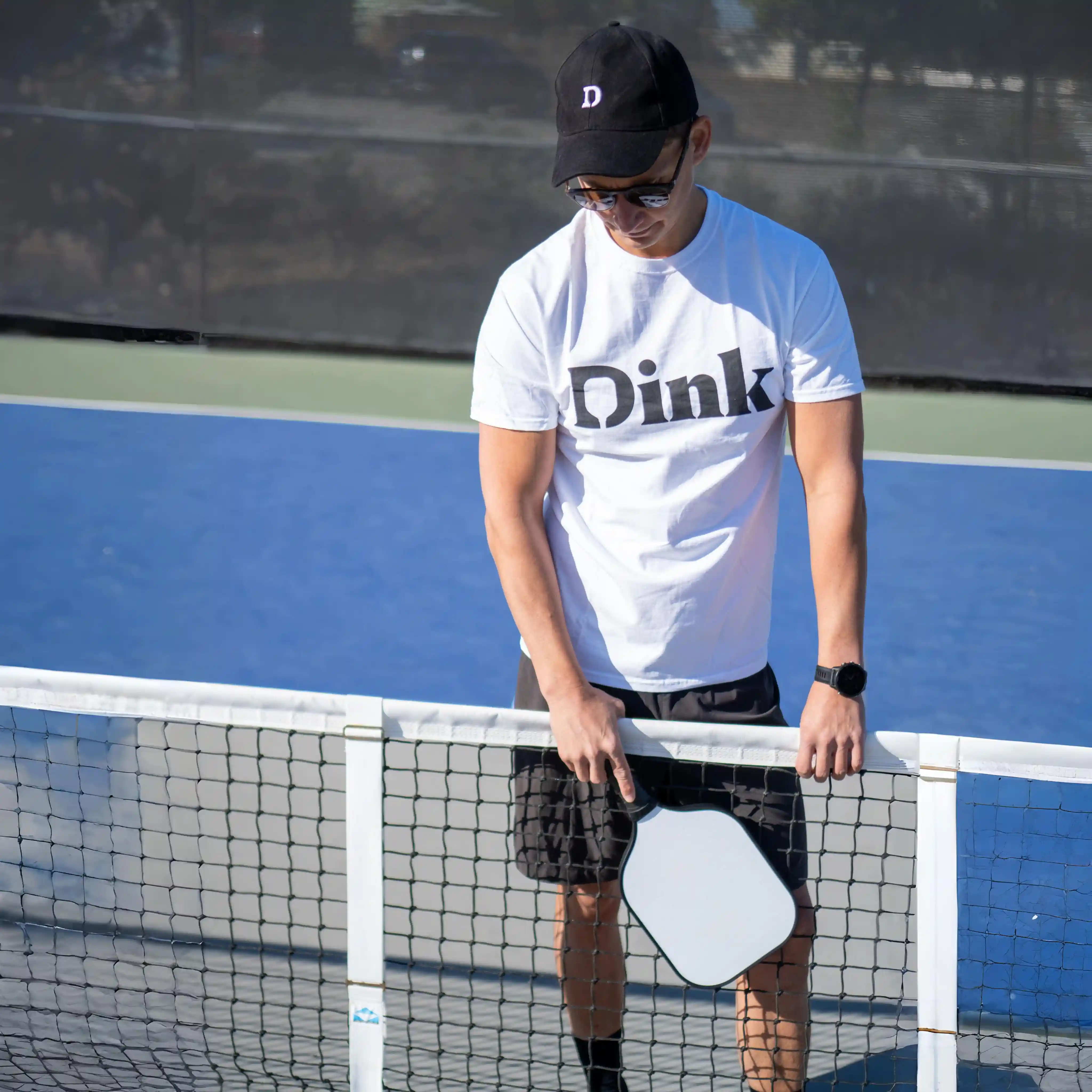 Man in a 'Dink' shirt holding a pickleball paddle on a court